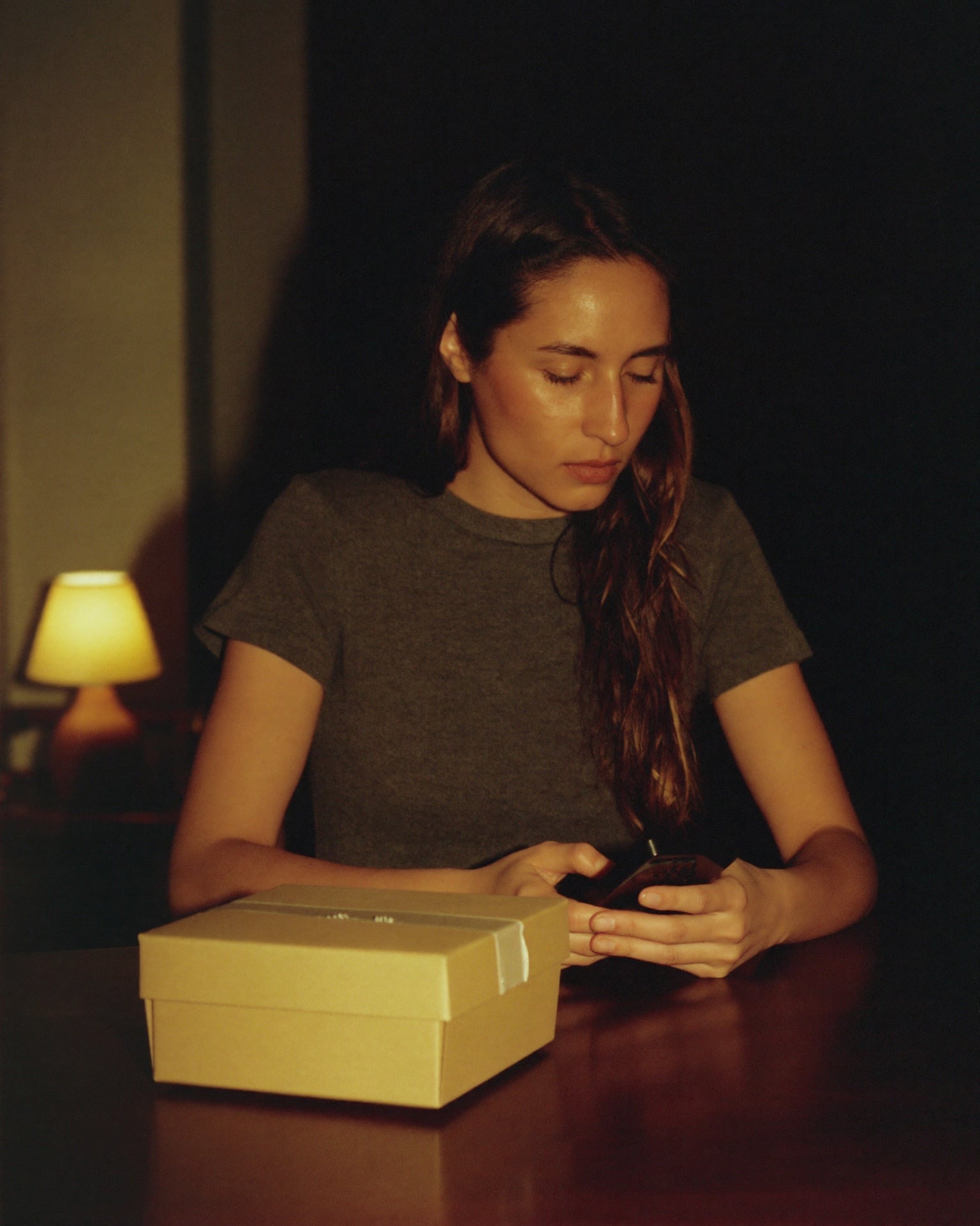 A woman sits at her own table in lamplight late in the evening, phone held low in both hands, a plain unmarked parcel on the table in front of her